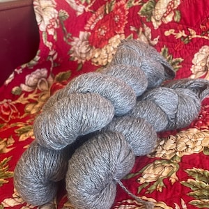 May include: Several skeins of gray yarn are arranged on a red floral patterned fabric. The yarn appears to be soft and fluffy, ideal for knitting or crocheting projects. The background fabric has a vintage aesthetic.