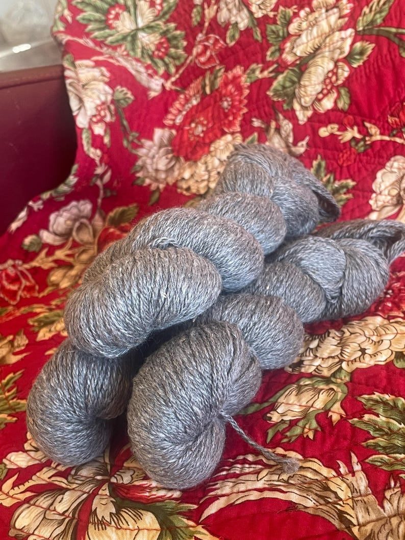 May include: Several skeins of gray yarn are arranged on a red floral patterned fabric. The yarn appears to be soft and fluffy, ideal for knitting or crocheting projects. The background fabric has a vintage aesthetic.