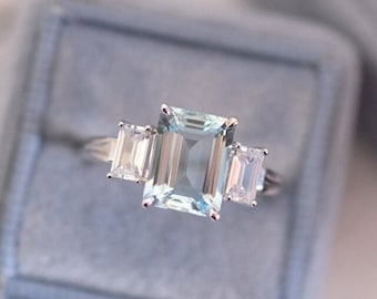 May include: A three-stone engagement ring with a large, emerald-cut aquamarine center stone and two smaller, baguette-cut diamond side stones. The ring is set in a white metal band.