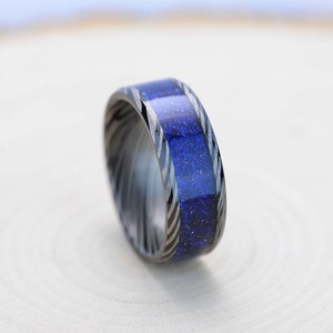 Pode incluir: Um anel com uma banda de metal escura e estampada e uma pedra central azul brilhante. A pedra azul tem uma aparência salpicada. O anel está sobre uma superfície de madeira clara.