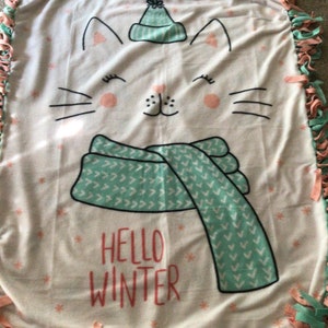 May include: White fleece blanket with a pink and green knotted fringe border. The blanket features a cartoon cat wearing a green hat and scarf. The text "Hello Winter" is printed on the blanket.
