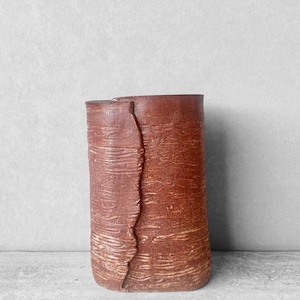 May include: A handmade, cylindrical ceramic vase in a reddish-brown hue. The vase features a textured surface with horizontal lines and a vertical, jagged split down the side. The design gives the vase a rustic, artisanal aesthetic.