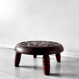 May include: A round, dark brown wooden stool featuring a carved geometric star pattern on its flat top surface. The stool is supported by four short, turned legs that provide a low profile, showcasing a smooth, polished finish throughout.