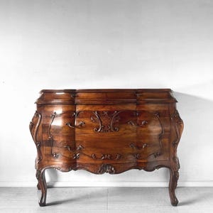May include: A dark brown wooden chest of drawers featuring a curved front, three horizontal drawers, and intricate carved detailing along the edges and drawer fronts. The piece stands on four curved legs and includes metal drawer pulls.