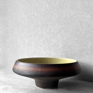 May include: A wide, shallow bowl with a dark brown, textured exterior featuring horizontal ridges and a smooth, pale yellow interior. The bowl has a tapered, circular base that elevates the main body, creating a distinct silhouette.