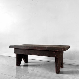 May include: A low, rectangular wooden bench featuring a dark, weathered brown finish with visible wood grain. The bench is constructed with a thick, flat top surface supported by four sturdy, angled legs, showcasing a simple, functional design.
