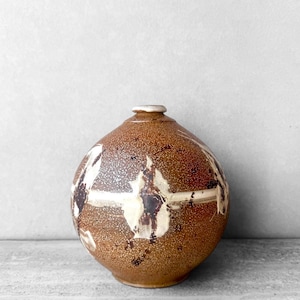 May include: A spherical ceramic vase with a textured brown speckled glaze. The surface features a horizontal band of white and dark brown abstract markings that wrap around the center of the rounded body, leading to a small, narrow neck opening.
