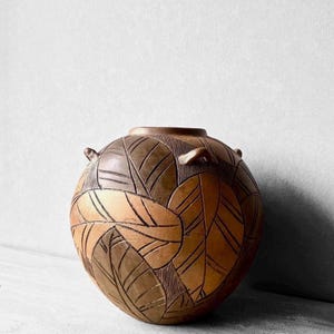 May include: A spherical ceramic vase featuring a two-tone brown and tan color palette. The surface is decorated with deeply incised leaf patterns and fine line textures. The vessel has a small circular opening and two small side handles.