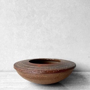 May include: A low-profile, circular ceramic bowl with a wide, flat rim. The exterior features a textured, speckled brown finish with subtle concentric circular grooves etched into the top surface, surrounding a deep, smooth, dark brown interior.