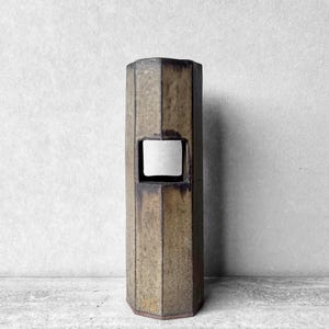 May include: A tall, geometric ceramic vase with a rectangular opening. The vase has a multi-sided shape and a textured, earthy brown and tan color scheme. The opening reveals a white interior.