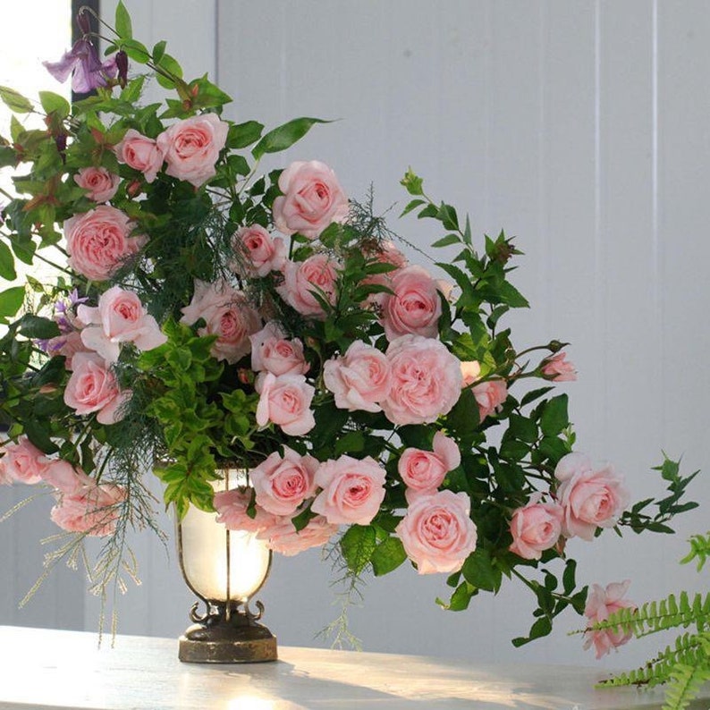May include: A bouquet of pink roses arranged in a clear glass vase with a gold metal base. The roses are surrounded by green leaves and stems.