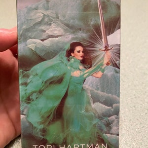 May include: A woman in a flowing green gown holds a silver sword in a snowy landscape. The text "TORI HARTMAN" and "Bestselling Author of the Chakra Wisdom Oracle Cards" is at the bottom of the image.