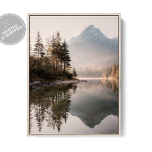 May include: A framed landscape print featuring a serene mountain lake scene. The image shows a mountain reflected in the calm water, with trees lining the shore. The print has a light-colored frame and a soft, natural color palette.
