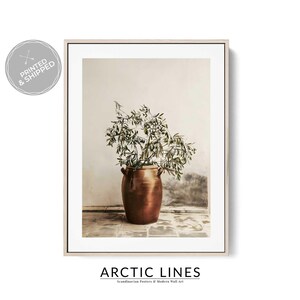 Op de afbeelding: Een ingelijste print met een bronzen vaas met groene, bladerrijke takken tegen een neutrale muur en een betegelde vloer. Het kunstwerk heeft de tekst "ARCTIC LINES" onderaan. De print zit in een lichtgekleurde lijst.