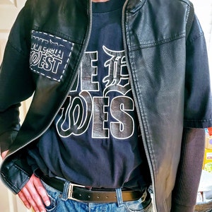 Könnte beinhalten: Eine schwarze Lederjacke mit Reißverschluss und darunter ein schwarzes T-Shirt. Das T-Shirt hat einen weißen Aufdruck mit der Aufschrift "Wild West" und einen kleineren Patch mit der Aufschrift "Wild West" auf der linken Brustseite.