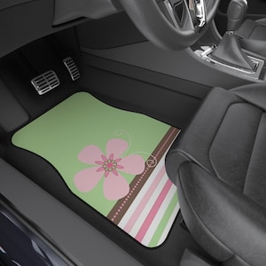 May include: A green car floor mat with a large pink flower design and a brown dotted stripe. The mat also features pink and white stripes. The mat is placed in the driver's side of a car.