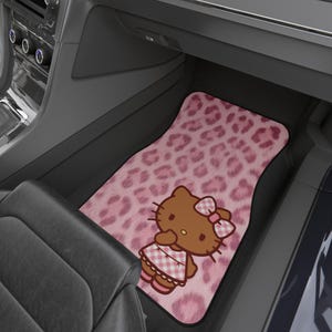 May include: A car floor mat featuring a pink leopard print background and a brown Hello Kitty character wearing a pink and white checkered dress and a matching bow. The character is positioned in the lower center of the rectangular mat.