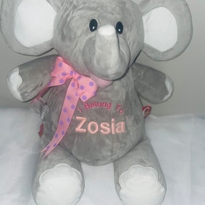 May include: A plush grey elephant toy with large ears featuring white inner panels. The elephant has white foot pads, black eyes, and a pink polka-dot ribbon tied around its neck. Embroidered in pink on the chest is the text I Belong To Zosia.