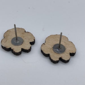 May include: A pair of wooden flower-shaped earrings with silver posts and backs. The earrings are a light brown color and have a natural wood grain.