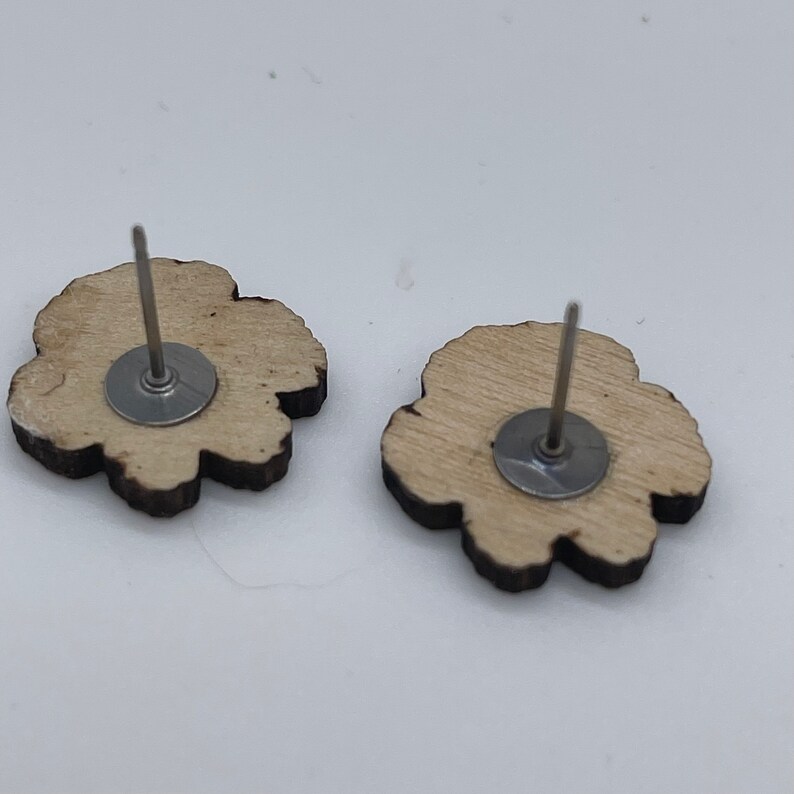May include: A pair of wooden flower-shaped earrings with silver posts and backs. The earrings are a light brown color and have a natural wood grain.