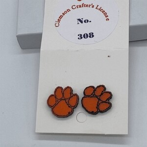 May include: A pair of orange and black paw print earrings with a white card behind them. The card has the text "Clemson Crafter's License No. 308" printed on it.