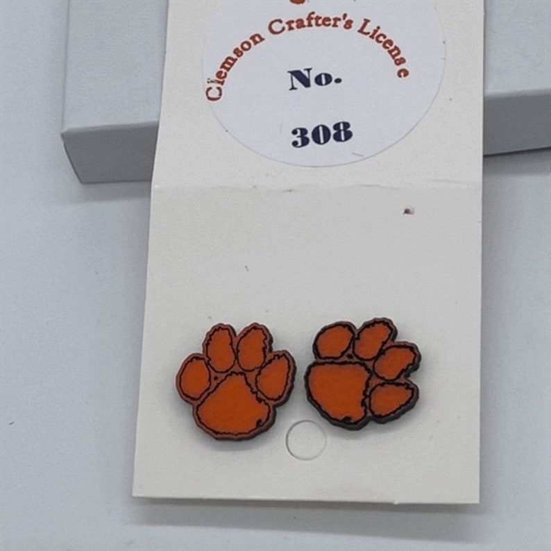 May include: A pair of orange and black paw print earrings with a white card behind them. The card has the text "Clemson Crafter's License No. 308" printed on it.