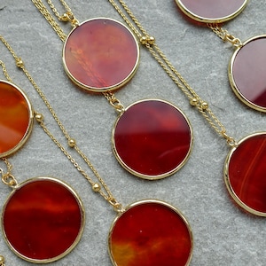 May include: Gold necklaces featuring circular, red-orange pendants. The pendants have a gold border and hang from fine gold chains. The necklaces are displayed on a grey surface, highlighting the jewellery's design and colour variations.