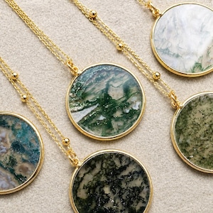 May include: Gold-toned necklaces with round pendants. The pendants feature natural stone with green, white, and black patterns. The chains have small gold beads. The necklaces are displayed on a neutral surface.