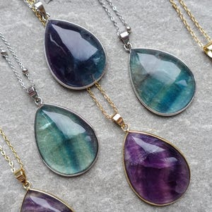 May include: Close-up of several gemstone pendant necklaces. The pendants are teardrop-shaped, with a mix of purple, blue, and green hues. Some have silver chains, while others have gold chains. The necklaces are displayed on a grey surface.