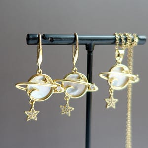 May include: A gold-coloured jewellery set with a planet design, featuring a white centre and a sparkling star charm. The set includes drop earrings and a necklace with a delicate chain, displayed on a black stand.