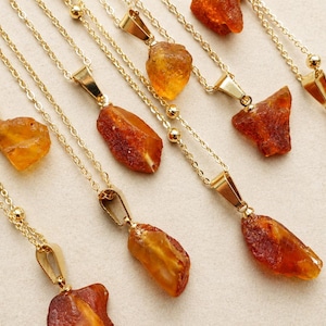 May include: A selection of gold necklaces, each featuring a raw amber pendant. The amber stones are in shades of orange and brown. The necklaces are displayed on a light-coloured surface.