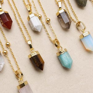 Op de afbeelding: Een verzameling gouden kettingen met verschillende edelsteenhangers. De hangers zijn in tinten rood, wit, bruin, grijs en blauw. Elke ketting heeft een fijne ketting en een kristallen hanger.