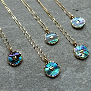 May include: A collection of gold necklaces, each featuring a pendant made from iridescent abalone shell. The pendants are adorned with a small gold heart. The necklaces are arranged on a grey surface.