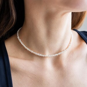 May include: A fine pearl necklace. The necklace features small, round, white pearls. The pearls are strung together to form a choker. The necklace is worn around the neck. The background is a dark blue top.