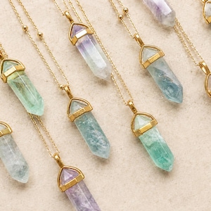 May include: A collection of necklaces featuring pointed crystal pendants in shades of green, purple, and clear, each encased in a gold-toned metal cap and suspended from a delicate gold-toned chain with small bead accents along the length.