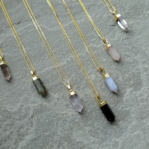 Könnte beinhalten: Eine Sammlung goldfarbener Halsketten, jede mit einem anderen Edelsteinanhänger. Die Anhänger sind in verschiedenen Farben erhältlich, darunter klar, rosa und schwarz. Die Halsketten werden auf einer grauen Oberfläche präsentiert.
