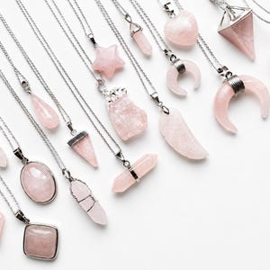 Puede incluir: Una colección de collares plateados con diversos colgantes de cuarzo rosa en formas que incluyen estrellas, lunas crecientes, corazones, pirámides, puntas y formas geométricas, suspendidos en delicadas cadenas de metal plateado.
