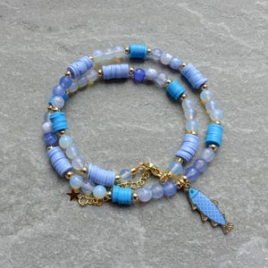 May include: A layered necklace featuring blue and white beads, with gold accents and a fish charm. The necklace has a mix of round and cylindrical beads in shades of blue and white, connected by a gold chain.