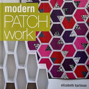 May include: A book titled "modern PATCH WORK" with a lime green cover. The book is next to a colorful quilt with a geometric pattern of hexagons in shades of red, pink, and purple. The quilt has black and white triangle accents.