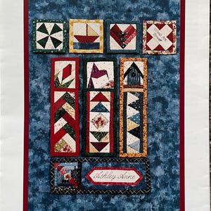 May include: A collection of handmade fabric bookmarks with various designs, including a sailboat, hearts, and geometric patterns. The text "QUICK AS CAN BEE 2! BOOKMARKS" is at the top, with "Ashley Anne" in a banner. The background is a blue fabric.