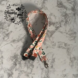 May include: A white lanyard with a floral pattern in shades of pink, green, and yellow. The lanyard has a black clip on one end and a silver swivel hook on the other. The lanyard is labeled "The Hunny Pot Shop".