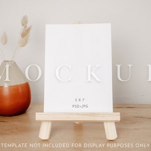 Op de afbeelding: Een witte 5x7 inch kaart mockup op een houten ezel. De kaart is leeg en klaar voor uw ontwerp. De ezel staat op een houten tafel.