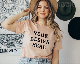 Può includere: Una maglietta color pesca chiaro con la scritta "YOUR DESIGN HERE" in grassetto nero. La modella indossa un cappello color cachi e jeans blu. Lo sfondo comprende un arazzo da parete con un disegno di sole e tre cappelli.
