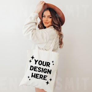 Peut inclure: Un sac fourre-tout en toile blanche avec du texte noir qui dit "YOUR DESIGN HERE" et de petites étoiles autour du texte. Le sac est tenu par une personne portant un pull blanc et un chapeau marron.