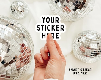 May include: A hand holding a white sticker with the text "YOUR STICKER HERE" in black letters. The sticker is in front of a white background with several silver disco balls.