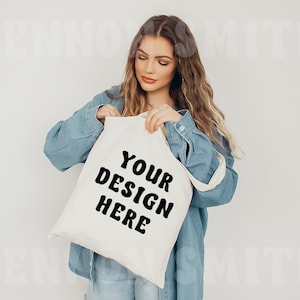 May include: A white canvas tote bag with black text that reads "YOUR DESIGN HERE". The bag is being held by a person wearing a blue denim shirt.