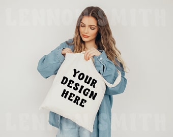May include: A white canvas tote bag with black text that reads "YOUR DESIGN HERE". The bag is being held by a person wearing a blue denim shirt.