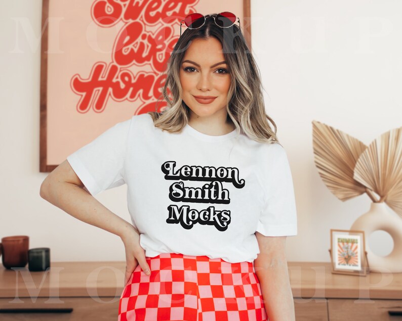 Puede incluir: Una camiseta blanca con texto negro que dice "Lennon Smith Mocks". La modelo lleva la camiseta con una falda de cuadros rojos y blancos y gafas de sol.