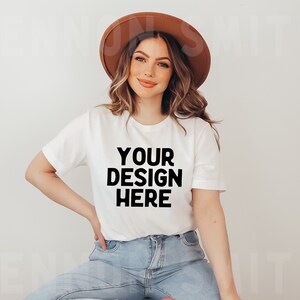 May include: A white t-shirt with the text "YOUR DESIGN HERE" printed in black. The shirt is being worn by a person with long brown hair and a brown hat.