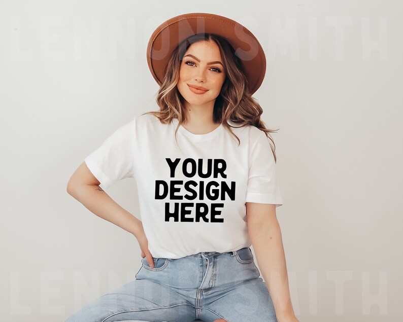May include: A white t-shirt with the text "YOUR DESIGN HERE" printed in black. The shirt is being worn by a person with long brown hair and a brown hat.